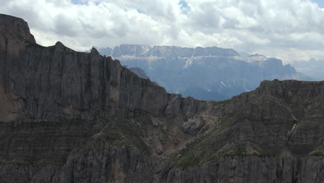 majestic aerial footage of towering rocky cliffs with distant sellaronda snow-capped mountain ranges in the dolomites, italy, under a partly cloudy sky