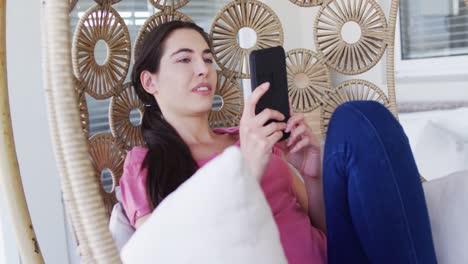 Happy-caucasian-woman-sitting-on-hanging-chair-on-balcony-and-using-smartphone