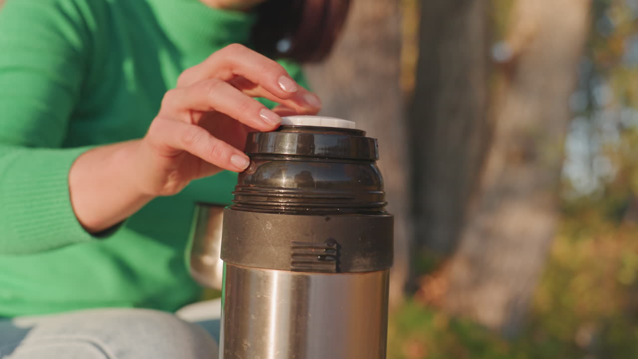 White woman camping pours coffee outdoors. Relaxed camper sits in folding chair near hammock, opens stainless thermos, pours hot coffee into mug, warm sun through autumn trees, cooler and gear nearby