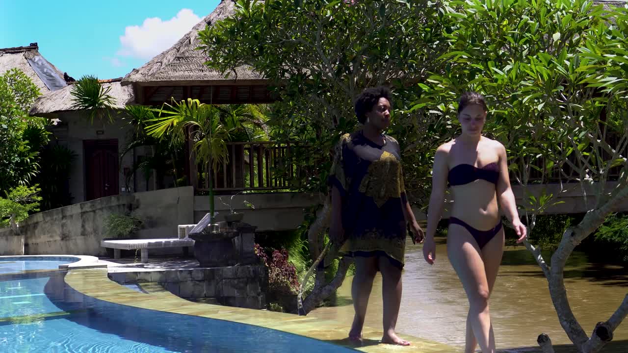 two young women walking casually along the swimming pool in luxury hotel Santi Mandala.  wearing fancy bikini and doing the cat walk along the pool