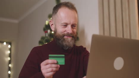 vista inferior de un hombre de cabello rojo que compra en línea con una tarjeta de crédito usando una computadora portátil sentada en una mesa en una habitación decorada con un árbol de navidad 1