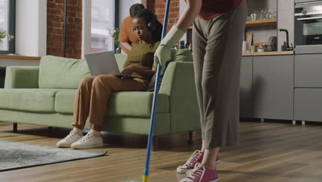 Afroamerikanisches-Mädchen-Mit-Laptop-Auf-Sofa-Sitzend,-Während-Mitbewohner-Das-Haus-Putzen-1