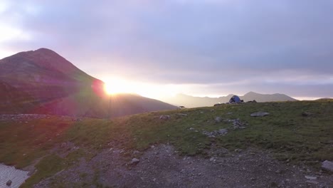 descending drone view at sunset with cloudy sky and tent on ridge with view of negoiu in the distance as sun sets behind the far ridge