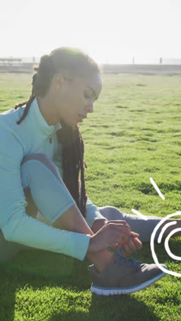 Sports-concept-icons-against-african-american-fit-woman-tying-her-shoe-laces-sitting-on-the-grass