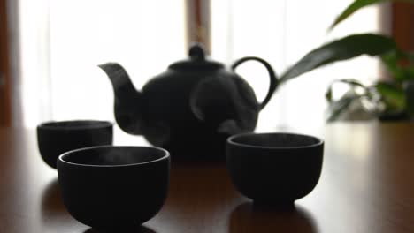 minimal background of a green japanese tea set with steam coming out of the cups, on a wooden table, with a plant and a window in the back