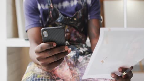 Midsection-of-african-american-male-painter-holding-smartphone-and-photograph-in-artist-studio