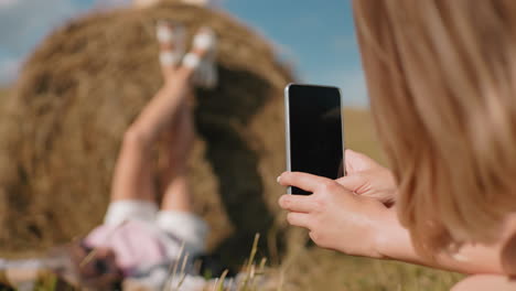 mujer tomando una foto con un teléfono inteligente de un amigo acostado en el heno con las piernas levantadas en el aire, un entorno al aire libre soleado en un campo rural, capturando un momento divertido y relajado en la naturaleza