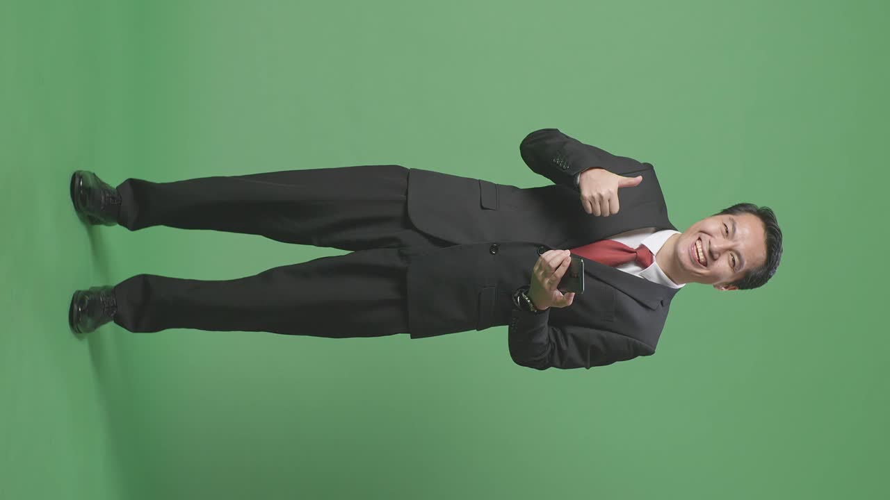 스마트폰을 사용하는 아시아 사업가의 몸이 가득 차 있고 스튜디오에서 녹색 화면에 서서 엄지손가락을 보여주고 있습니다.