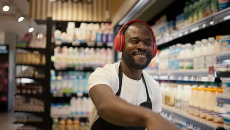 Un-Hombre-De-Piel-Negra-Con-Auriculares-Rojos-Y-Un-Delantal-Negro-Baila-En-El-Departamento-De-Lácteos-De-Un-Supermercado.-Feliz-En-El-Trabajo