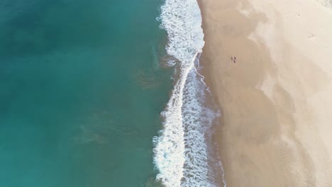 Luftaufnahme-Der-Wellen-Des-Blauen-Meeres,-Die-Sich-Auf-Dem-Sand-Eines-Wunderschönen-Langen-Weißen-Sandstrandes-Und-Einiger-Weißer-Häuser-In-Der-Ferne-Brechen