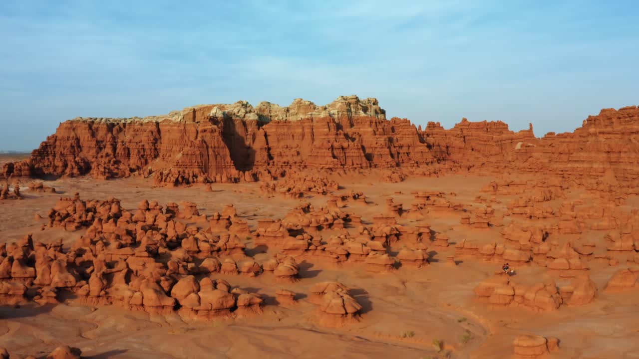 Imponerende droneoptagelse fra luften, der bevæger sig til venstre, af den smukke Goblin Valley Utah State Park med små, mærkelige svampelignende klippeformationer nedenfor og store røde og hvide buttes i baggrunden
