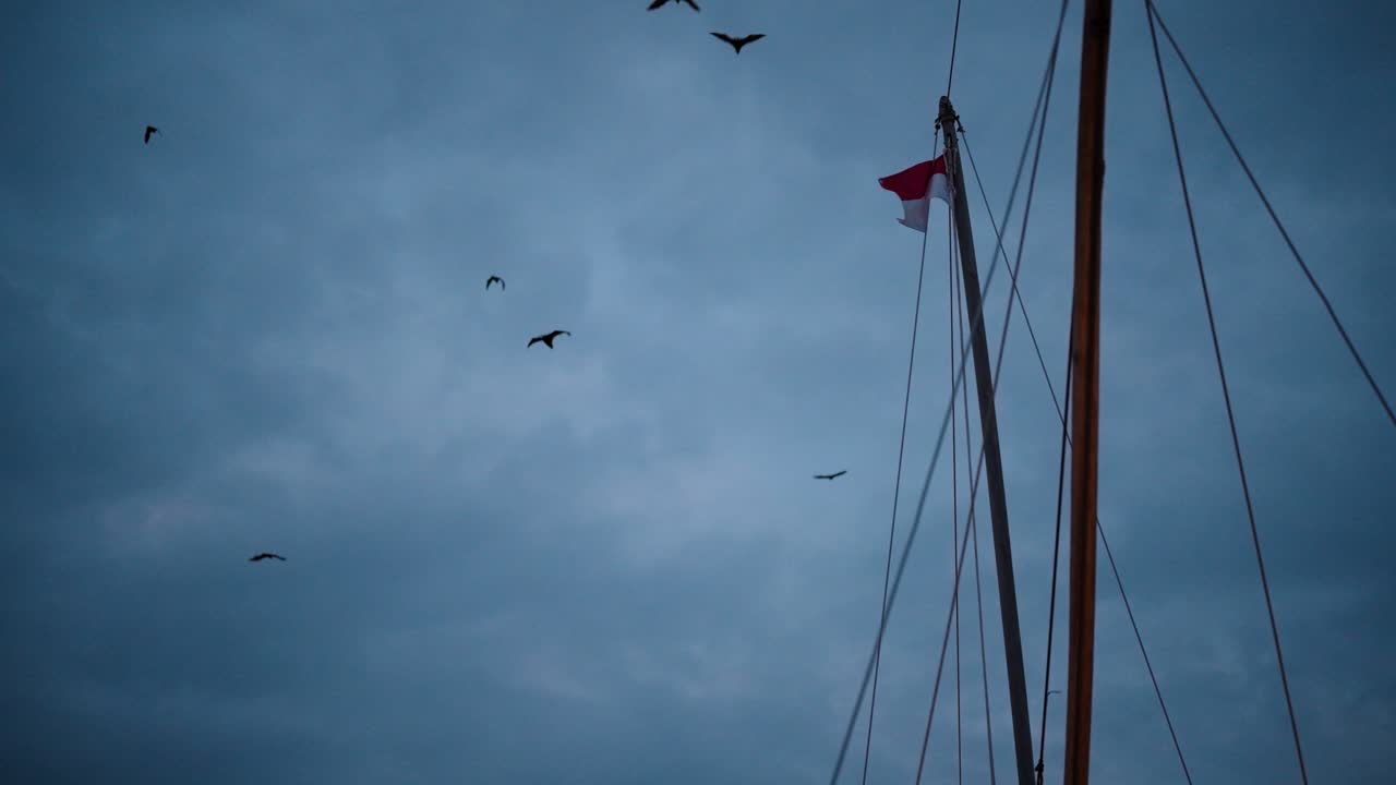 Fruit Bats also known as Flying Foxes fly over a boat with the mast and an Indonesian flag showing against a cloudy sky in the Komodo National Park