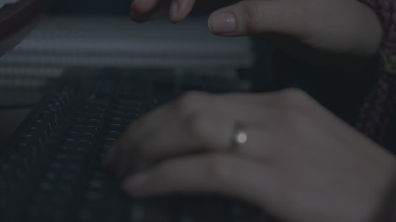 Primo piano di mani femminili che digitano su una tastiera in una stanza scarsamente illuminata. La scena trasmette concentrazione e attenzione. La luce soffusa aggiunge un'atmosfera suggestiva, evidenziando l'azione di digitare.