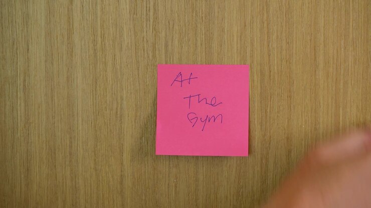 Close-up shot of a hand placing a bright pink sticky note reading 'at the Gym' on a textured wooden door, in soft lighting. The focus is on texture and color contrast