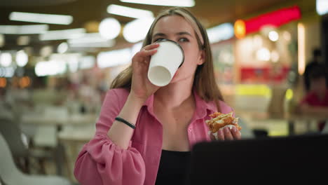 woman in pink dress taking a sip of coffee while holding a burger in her left hand, sitting in a mall restaurant, she continues working after taking a drink, with a busy, blurred background