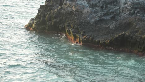 View-of-calm-ocean-surface-gently-hitting-on-rugged-side-of-cliff,-closeup