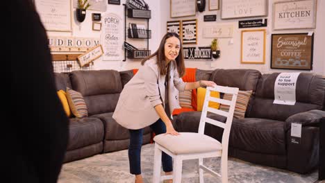 a young professional commission associate woman in business clothing stands over a wooden dining chair inside a retail furniture store workplace learning product knowledge from her sales manager