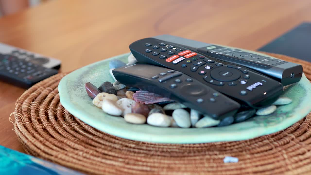 A hand methodically places multiple remote controls onto a decorative plate filled with pebbles and seashells, in a brightly lit home setting