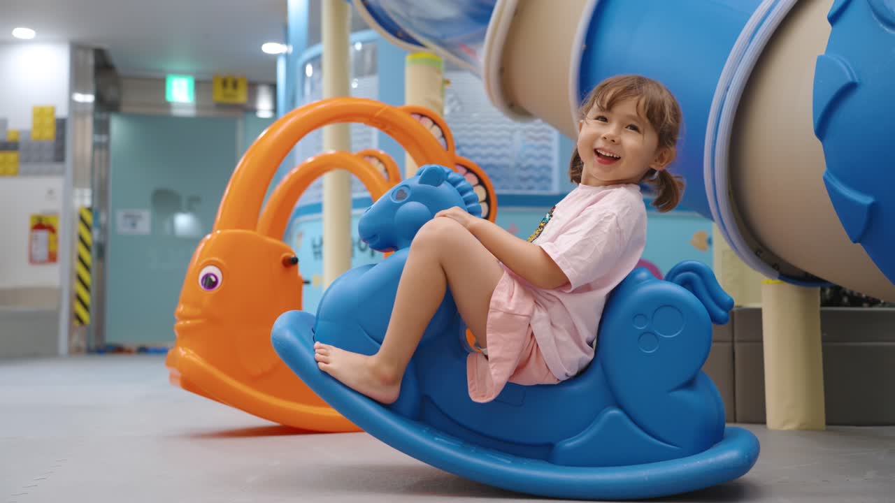 Young girl riding and rocking on a big colorful indoor playground blue toy horse, enjoying the fun ride with a bright vibrant smile, sideview medium static, slow motion