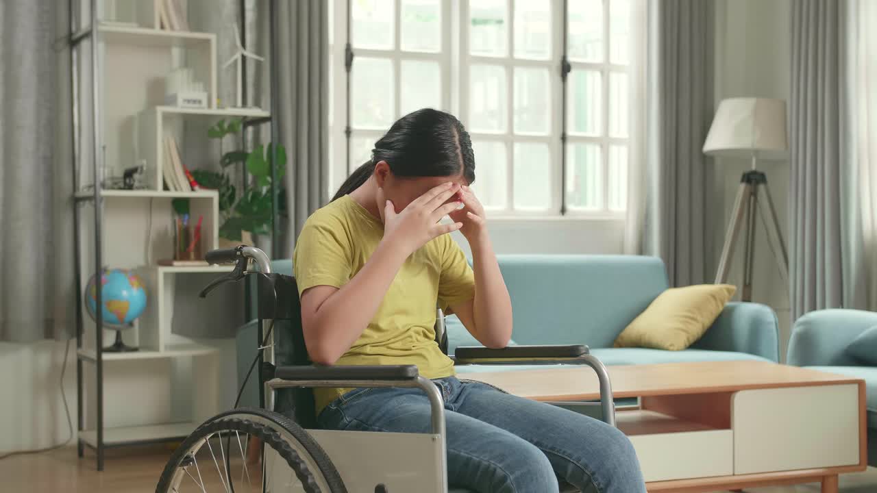 Disappointed Asian Kid Girl Sitting In A Wheelchair In Living Room At The House