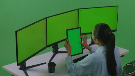 vista lateral de una mujer asiática trabajando con una tableta y varios monitores de computadora con una maqueta de pantalla verde en un estudio de pantalla verde