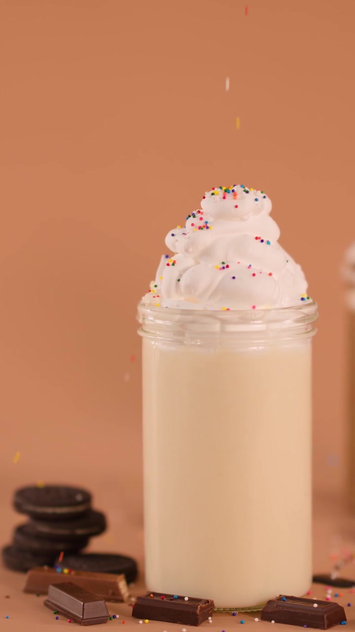 Granella che cade al rallentatore su un milkshake con panna montata in un barattolo di vetro, circondato da biscotti e cioccolato.