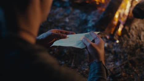 a person's hands interacting with a paper by a campfire at night