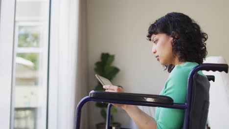 Thoughtful-biracial-disabled-woman-in-wheelchair-using-tablet-in-living-room
