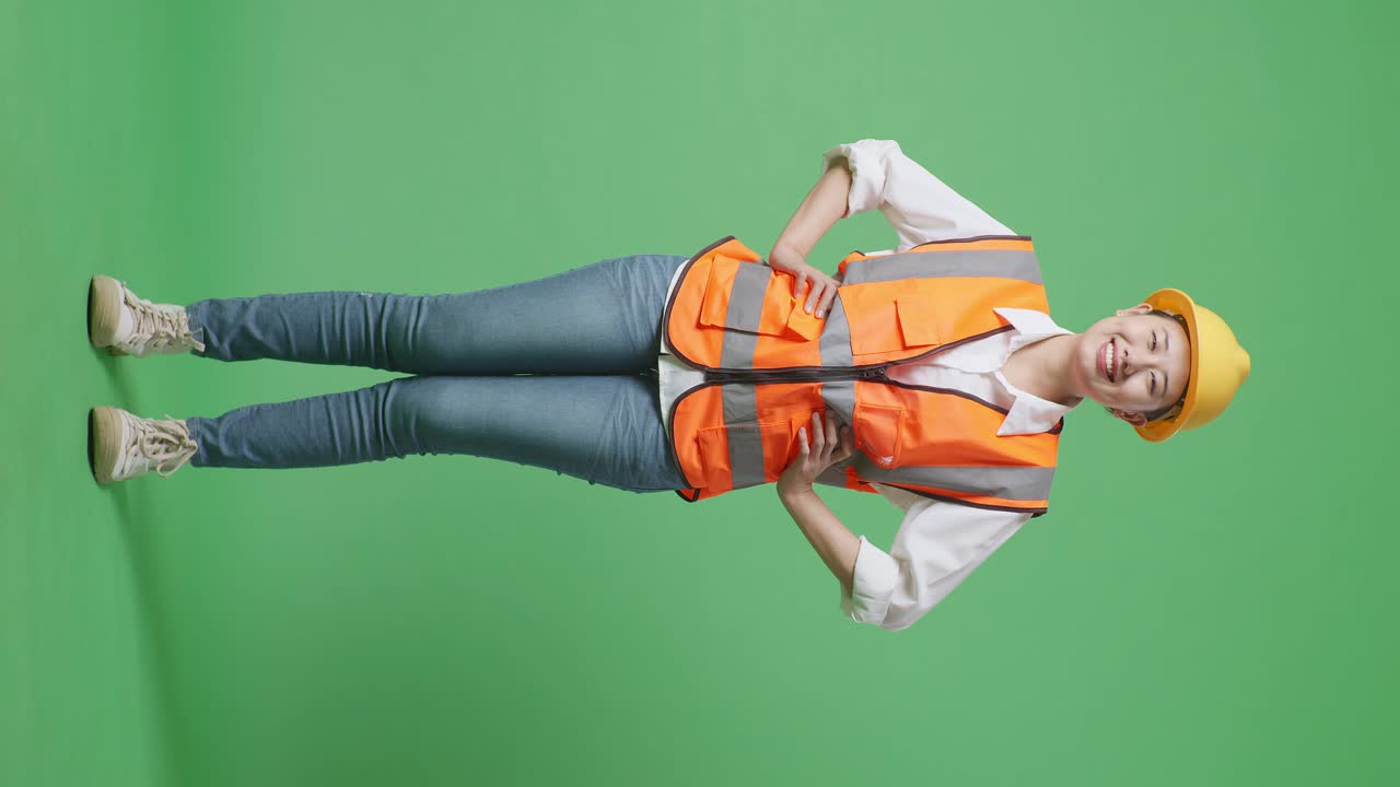 Full Body Of Asian Female Engineer Wearing Safety Helmet Looking At Camera While Standing With Arms Akimbo In The Green Screen Background Studio