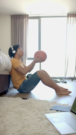 vertical video: listening to music with headphones, asian teenage boy sitting on floor tossing baske