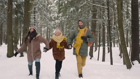 vista frontal de padres e hija vestidos con ropa de invierno caminando y saltando en un bosque nevado