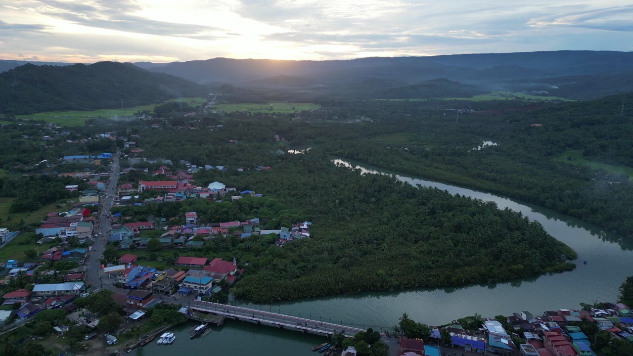 A forward sky view drone shot over a colorful riverside town with boats, a central bridge, lush green hills, and a warm sunrise glow over the winding river of Mauban Port, Quezon Province Philippines