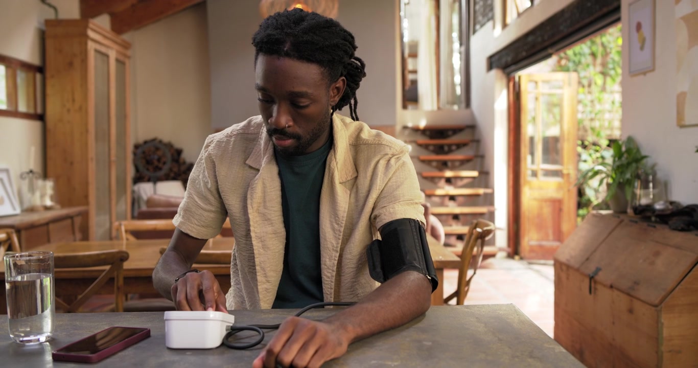 African American man rolling sleeve fitting cuff joining tubing to white unit at home monitoring BP
