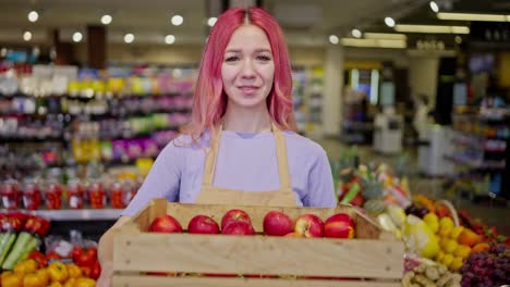 Porträt-Eines-Mädchens-Mit-Rosa-Haaren-In-Einer-Schürze,-Das-Mit-Einer-Kiste-Äpfel-Posiert,-Während-Es-In-Einem-Supermarkt-Arbeitet