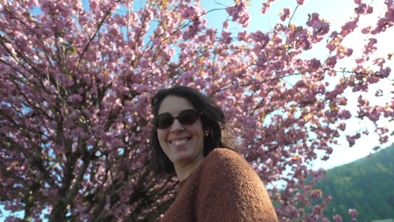 Mulher sorrindo com óculos de sol tendo como fundo uma árvore de cerejeira em flor rosa