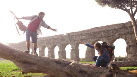 happy young couple backpackers tourists on a log trunk man with guitar reaching out with hand for woman in front of ancient roman aqueduct ruins in parco degli acquedotti park in rome at sunrise romantic slow motion