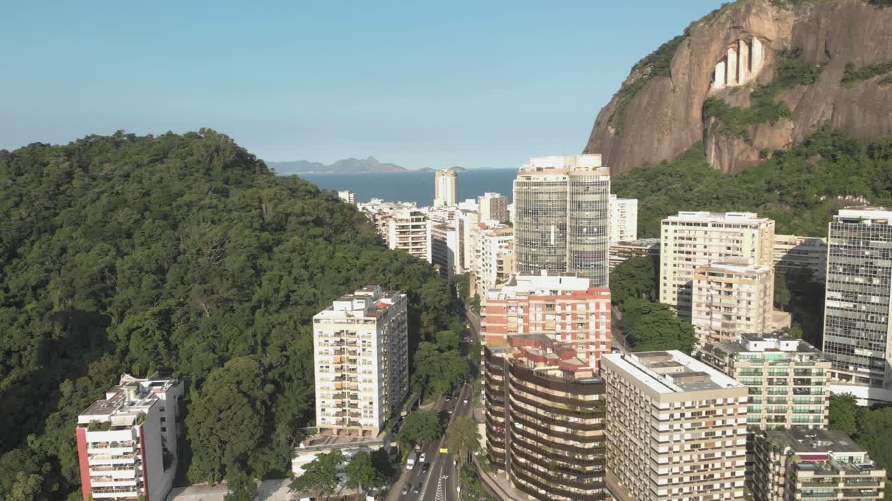 Movimento aéreo lateral lento para a esquerda com vista de edifícios residenciais altos com uma montanha ao fundo, revelando uma passagem em direção ao bairro de Copacabana no Rio de Janeiro.