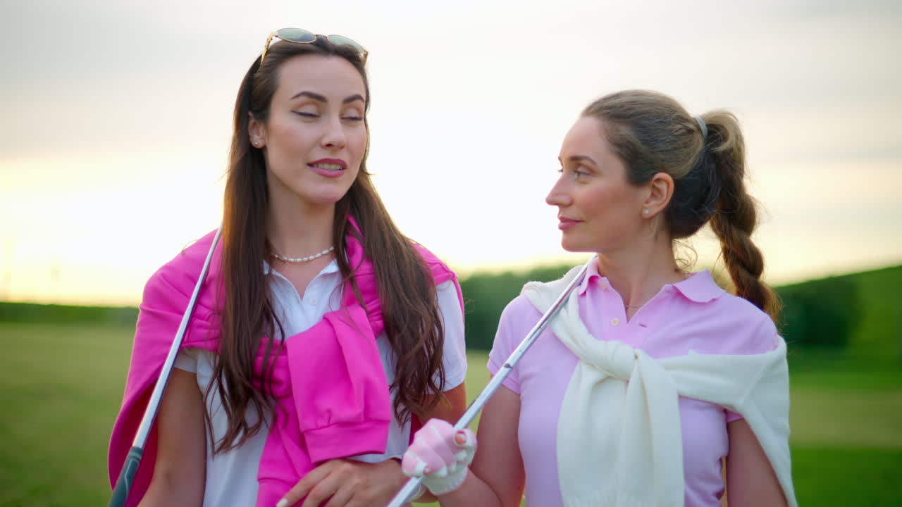 Two women dressed in white and pink clothes, with golf clubs on their shoulders, walking and talking on the golf course