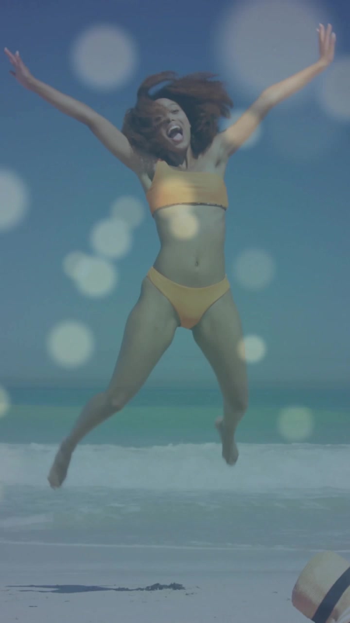 Video verticale: Donna che si accovaccia e poi si lancia in salti sulla spiaggia, con bokeh fluttuante, che evoca il viaggio.