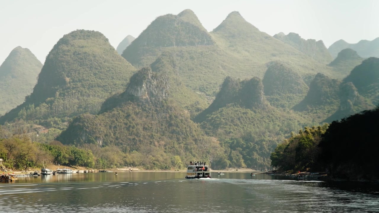 Thuyền du lịch di chuyển dọc theo một con sông yên ả, được bao quanh bởi những đỉnh núi đá vôi cao chót vót.