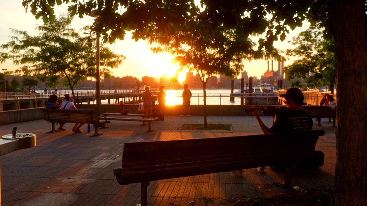 New York, USA, 5. Juni 2026: Geiger spielt am East River bei Sonnenuntergang in New York. Silhouettierter Geiger spielt auf einer Bank am East River mit Menschen und der Skyline von Manhattan bei Sonnenuntergang.