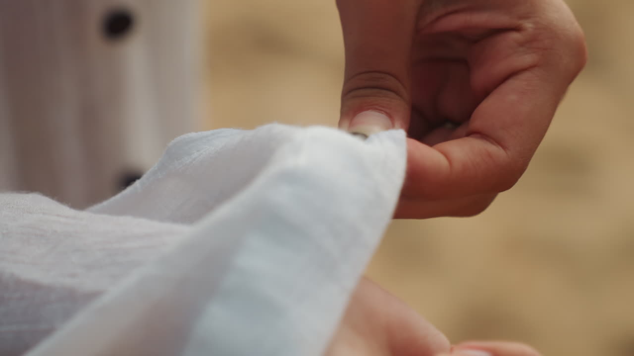 Primer plano de manos ajustando la manga de lino blanco en la playa durante la hora dorada, dedos doblando el puño para revelar pulsera y reloj, escenas de un suave ritual de preparación sugieren una novia arreglándose, un marinero revisando.