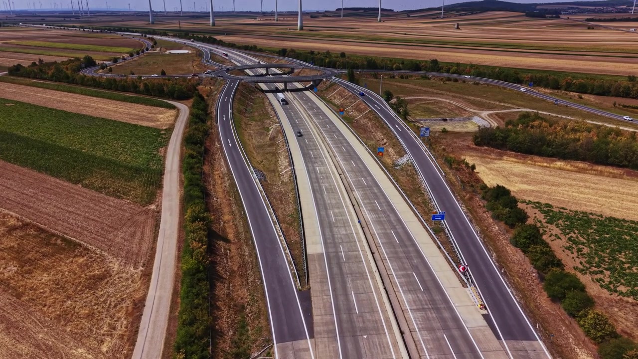 Veicoli su un'ampia autostrada circondata da campi coltivati e turbine eoliche.
