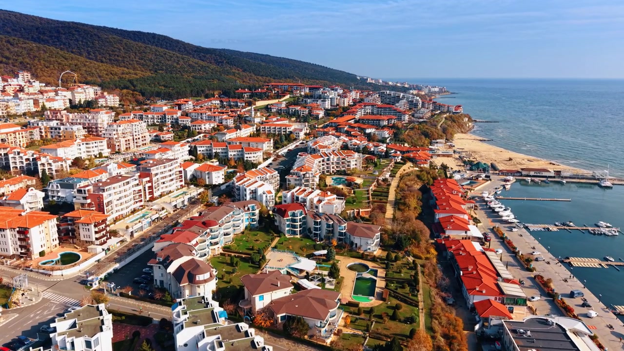 Vista elevata di un quartiere turistico costiero con porto turistico e colline boscose. Prospettiva aerea di edifici residenziali con tetti rossi che si estendono lungo la spiaggia e il porto con le barche.