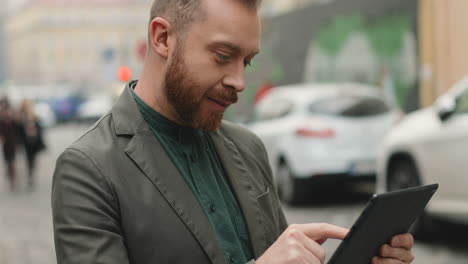 Close-up-view-of-smiled-young-Caucasian-businessman-standing-in-the-street-in-autumn-and-typing-on-the-tablet