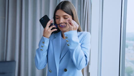 Serious beautiful young woman puts on glasses in office. Lady looks at her phone and then takes off her glasses and looks at window. Close up.