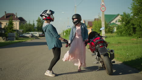 dos amigos con cascos de pie junto a una bicicleta eléctrica estacionada, compartiendo un momento alegre, una está jugando con su vestido rosa mientras la otra se ríe, postes eléctricos, casas y vegetación