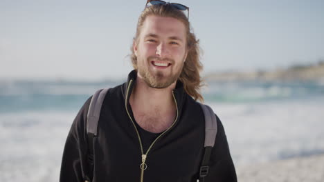 Retrato-De-Un-Joven-Apuesto-Sonriendo-Feliz-En-La-Playa-Disfrutando-De-Un-Cálido-Día-De-Vacaciones-De-Verano-En-La-Playa-Mirando-La-Cámara