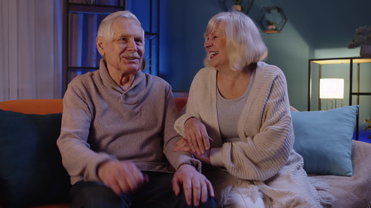 Happy old senior elderly family couple hugging, laughing, smiling talking at modern home sofa