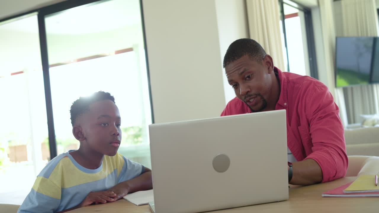 Papà afroamericano che usa il portatile mentre aiuta suo figlio a scrivere su un quaderno dopo aver alzato la matita al tavolo